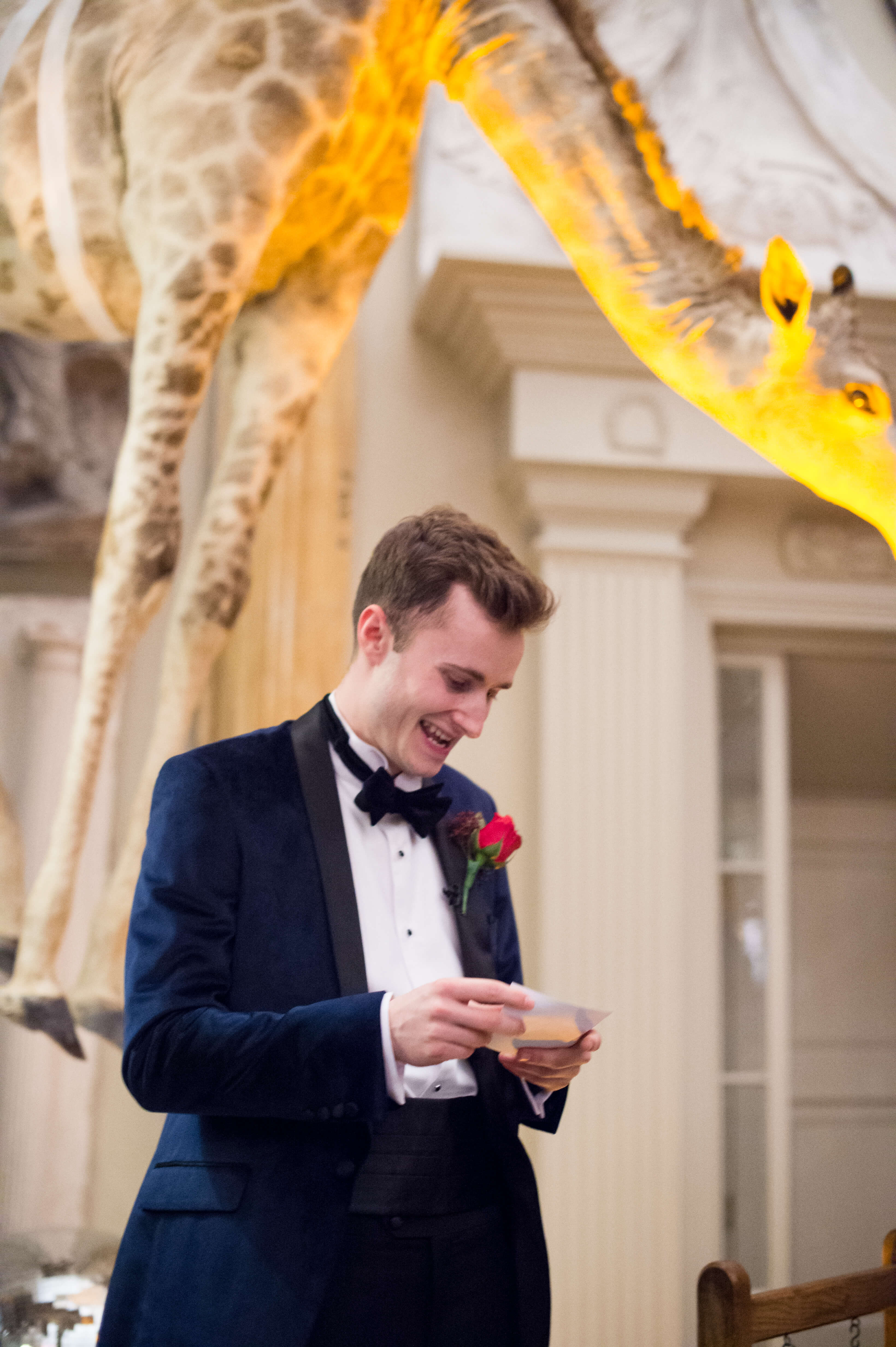 A photo of the best man giving a speech with a giraffe hanging behind him